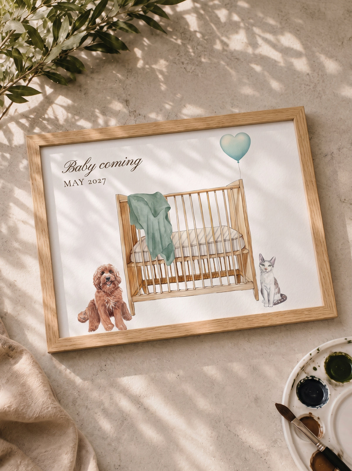 Pregnancy announcement watercolor: dog and cat beside a crib with a sage blanket and heart balloon, 'Baby coming May 2027', in an oak frame